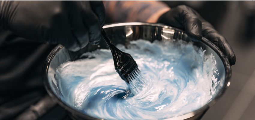 Gloved hands mixing vibrant blue bleach mixture in a bowl, demonstrating proper bleach to developer ratio.