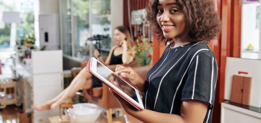 Happy customer using a tablet while waiting in a stylish lounge area of a franchise of salon, with another client receiving treatment in the background.