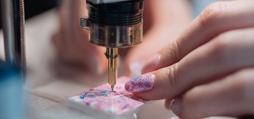 Close-up of hands with colorful spotted manicure pressing buttons on a modern nail printer, curing a fresh design under a UV/LED lamp for precise nail art printing.