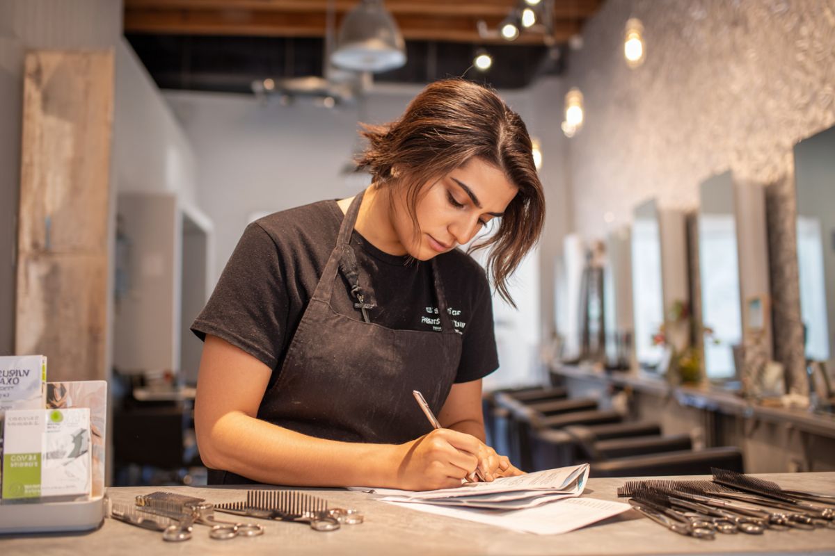 Focused hairstylist in black apron carefully filling out paperwork at her salon station, preparing documents related to her hairstylist license