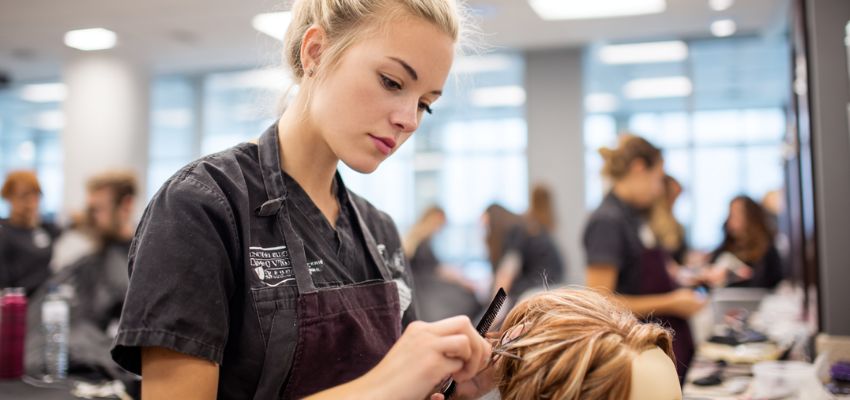 Professional hairstylist sectioning and cutting client's hair in a busy training salon, demonstrating skills required to earn a hairstylist license