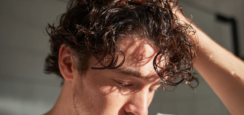 Close-up action shot of a man with type 2 hair during washing, wet dark brown wavy hair with pronounced S-shaped curls and foam, hand running through strands, water droplets visible, focused expression in a bathroom mirror setting