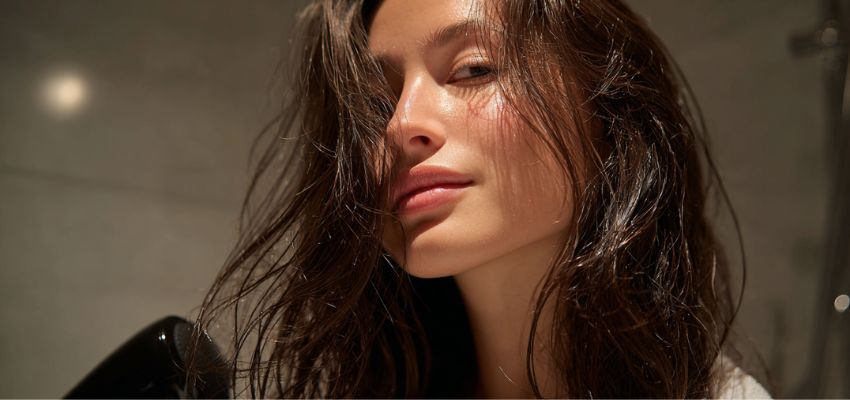 Woman with freshly washed wet hair in a steamy bathroom holding a blow dryer, capturing the moment people often wonder is blow drying hair bad for moisture and scalp health