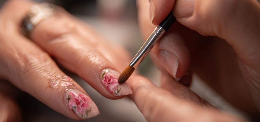 A detailed floral nail art design is carefully painted onto a fingernail with a fine brush during a manicure, illustrating how long does a manicure take.